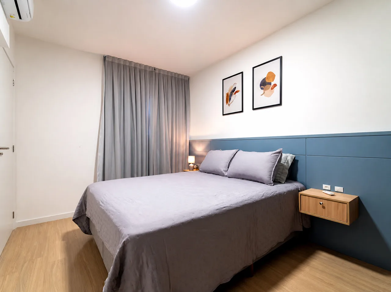 Quarto com cama queen, cabeceira azul estofada, criado-mudo suspenso e piso de madeira